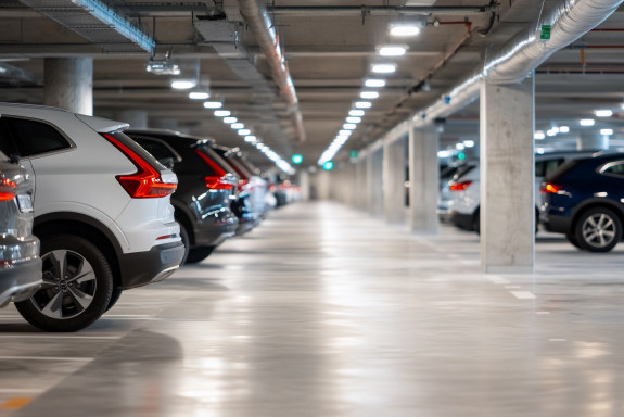 Rows of parked cars in a brightly lit underground garage with concrete pillars and ceiling pipes visible.