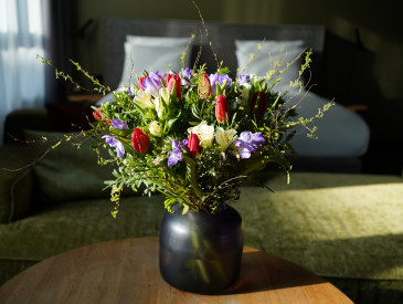 A bouquet of colorful flowers in a dark vase sits on a wooden table in a sunlit room with a bed and couch in the background.