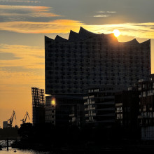 Die Sonne geht hinter einem modernen Glasgebäude am Wasser unter, in der Ferne sind Kräne zu sehen und über ihnen liegt ein goldener Himmel.