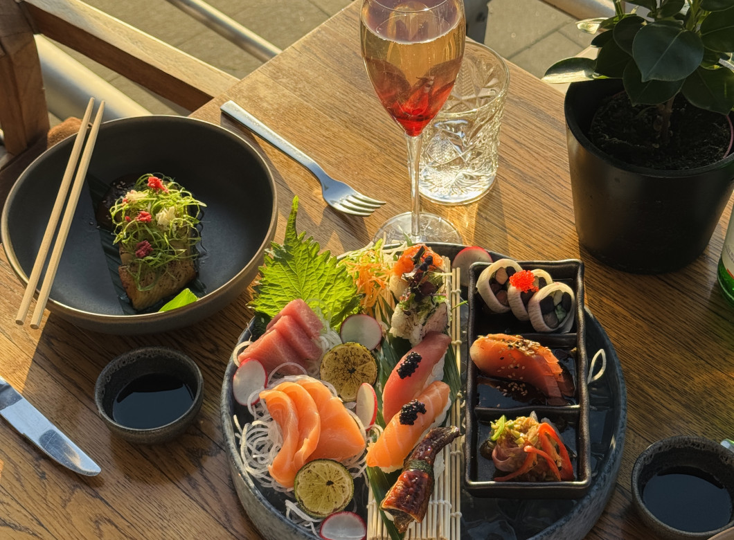 Ein Holztisch mit einer Auswahl an Sushi, Sashimi, Getränken und Besteck, in den Sonnenlicht einfällt und neben dem Essen Pflanzen stehen.