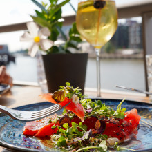 Auf einem blauen Teller liegt ein Stück Wassermelonensalat mit Grünzeug in einer Gabel, im Hintergrund ein Getränk und Blick auf den Fluss.