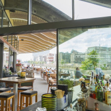 Ein modernes Restaurant am Flussufer mit Sitzgelegenheiten im Freien, Bartheke und Stadtgebäuden, die sich in großen Glasfenstern spiegeln.