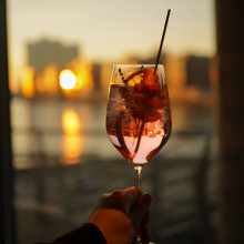 Eine Hand hält ein Glas rosa Cocktail mit Früchten und einem Strohhalm vor dem Hintergrund einer Stadtlandschaft bei Sonnenuntergang.